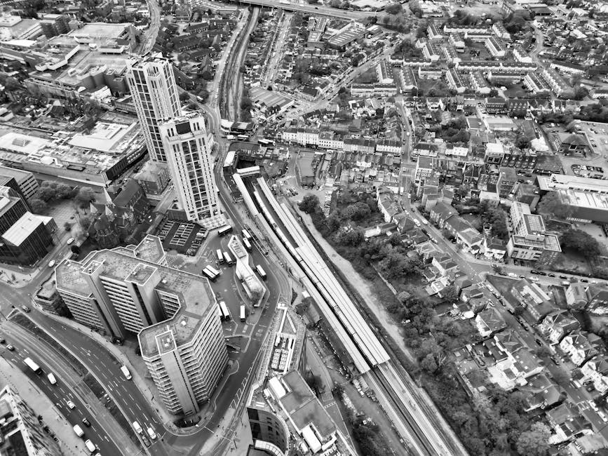 Aerial black and white photograph of an urban area showing a cluster of tall office buildings and high-rise apartments near Enfield Town station, with railway tracks running through the scene. The railway station platform is visible, with multiple trains parked and adjacent to a parking lot filled with vehicles. Surrounding the station, there are narrow streets lined with smaller residential houses and commercial buildings, with trees and greenery interspersed throughout the neighborhood. The image captures the dense layout of the city, emphasizing the proximity of residential, commercial, and transport infrastructure, which is relevant to home relocation and furniture transport services offered by Man with Van Enfield Town during moving and packing processes.