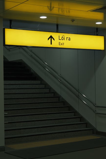A staircase inside an indoor transit area, with dark grey steps and metal handrails on both sides. Overhead, there is a bright yellow illuminated sign with black text and an upward-pointing arrow, indicating the exit, with the words 'Lối ra' and 'Exit' in both Vietnamese and English. The surrounding walls are smooth and light grey, and the lighting is even, providing clear visibility of the staircase and signage. This environment is typical of a train or bus station, where clear direction signs assist passengers in navigating toward exits, useful context for house removals or transport planning with companies like Man with Van Enfield Town that specialise in home relocation and furniture transport.