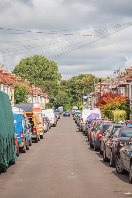 A delivery van with a white cab and an enclosed cargo area is parked on a narrow, sloped residential street in Enfield Town, surrounded by multi-storey brick and stucco buildings. The street is lined 