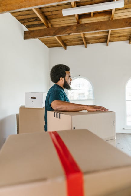 A man with dark curly hair and a beard is standing inside a bright, unfinished room with white walls and a wooden ceiling structure. He is wearing a teal t-shirt and is positioned behind several large cardboard moving boxes, one of which he has his hand resting on. The boxes are sealed with red packing tape and are stacked at waist height, likely prepared for a home relocation. The room has two arched windows allowing natural light to illuminate the space, and the floor appears to be wooden or laminate. In the background, the room is minimally furnished, and the ceiling structure is visible, indicating ongoing renovation or a move-in process. This scene exemplifies the packing and loading stages of house removals, with furniture and boxes ready for transport by [COMPANY_NAME], such as Man with Van Enfield Town, supporting efficient furniture transport and moving logistics.