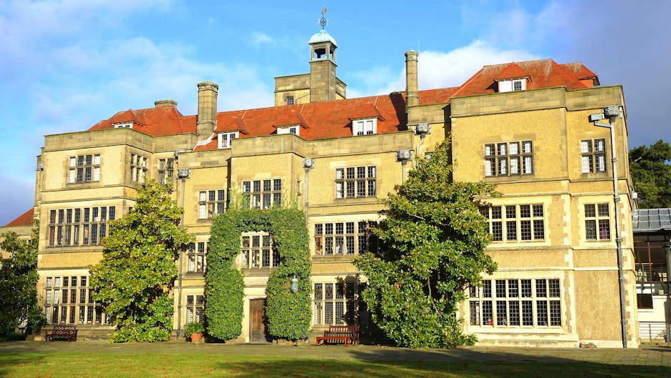 A large, historic building with a yellow stone facade, multiple multi-pane windows, and red-tiled roofs, situated in a landscaped setting with green trees and a well-maintained lawn in the foreground. The building features architectural details such as chimneys and decorative roof elements, with sunlight illuminating the structure against a partly cloudy sky. This image reflects the type of property often involved in house removals and furniture transport services provided by Man with Van Enfield Town, including packing, loading, and moving process visuals that relate to house relocation activities.