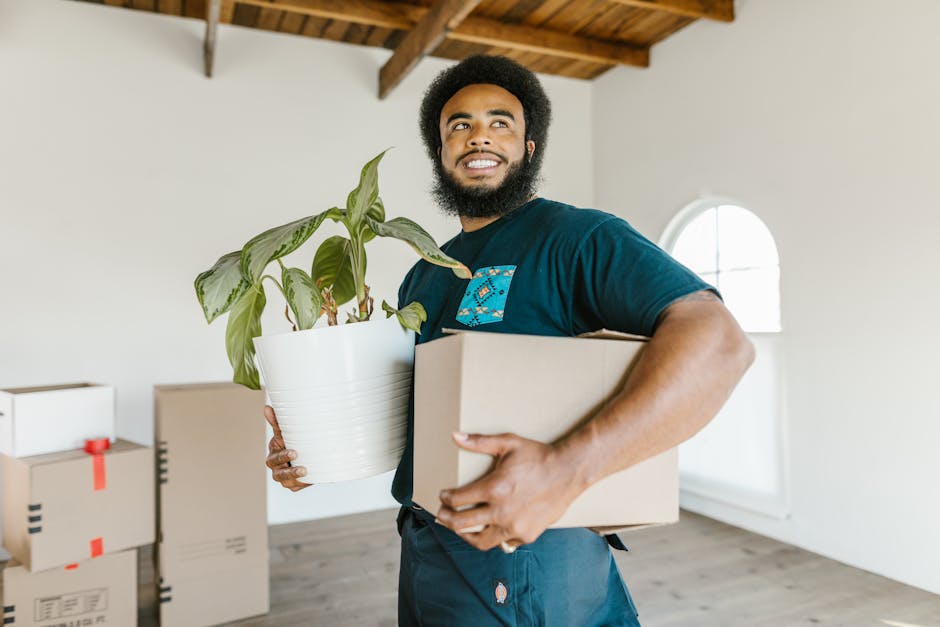 A man with dark curly hair and a beard is standing inside a bright, unfinished room with white walls and a wooden ceiling structure. He is wearing a teal t-shirt and is positioned behind several large cardboard moving boxes, one of which he has his hand resting on. The boxes are sealed with red packing tape and are stacked at waist height, likely prepared for a home relocation. The room has two arched windows allowing natural light to illuminate the space, and the floor appears to be wooden or laminate. In the background, the room is minimally furnished, and the ceiling structure is visible, indicating ongoing renovation or a move-in process. This scene exemplifies the packing and loading stages of house removals, with furniture and boxes ready for transport by [COMPANY_NAME], such as Man with Van Enfield Town, supporting efficient furniture transport and moving logistics.